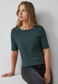 Top a maniche corte rigato di un verde profondo, scollatura rotonda, tessuto textured, design aderente, abbinato a jeans grigio scuro con finitura liscia.