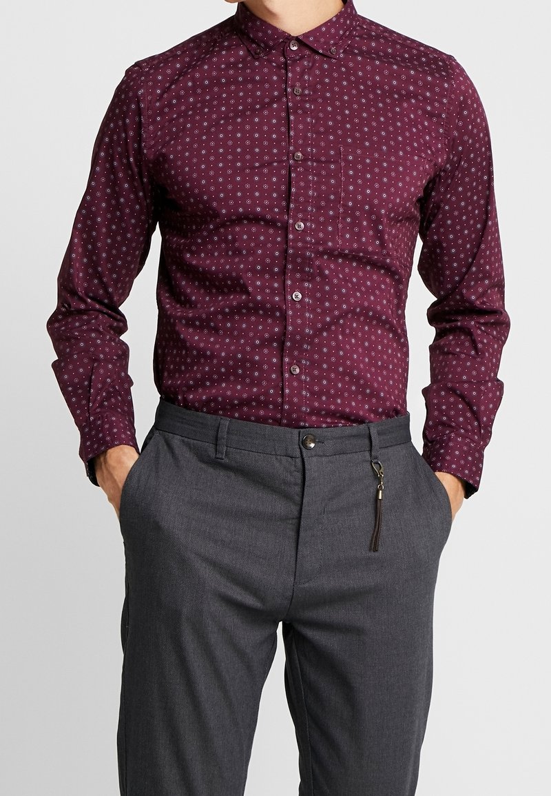 Homme portant une chemise bordeaux à manches longues avec de petits motifs circulaires blancs et un pantalon habillé gris, les mains dans les poches.