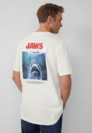 Crème katoen t-shirt met een grote grafische afbeelding op de achterkant van een haai, tekst met de woorden "JAWS" en een blauwe oceaan achtergrond.