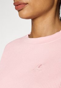 Sweatshirt rose clair avec une texture côtelée, dotée d'un col rond et d'un logo en relief sur la poitrine. Le tissu semble doux et flexible.
