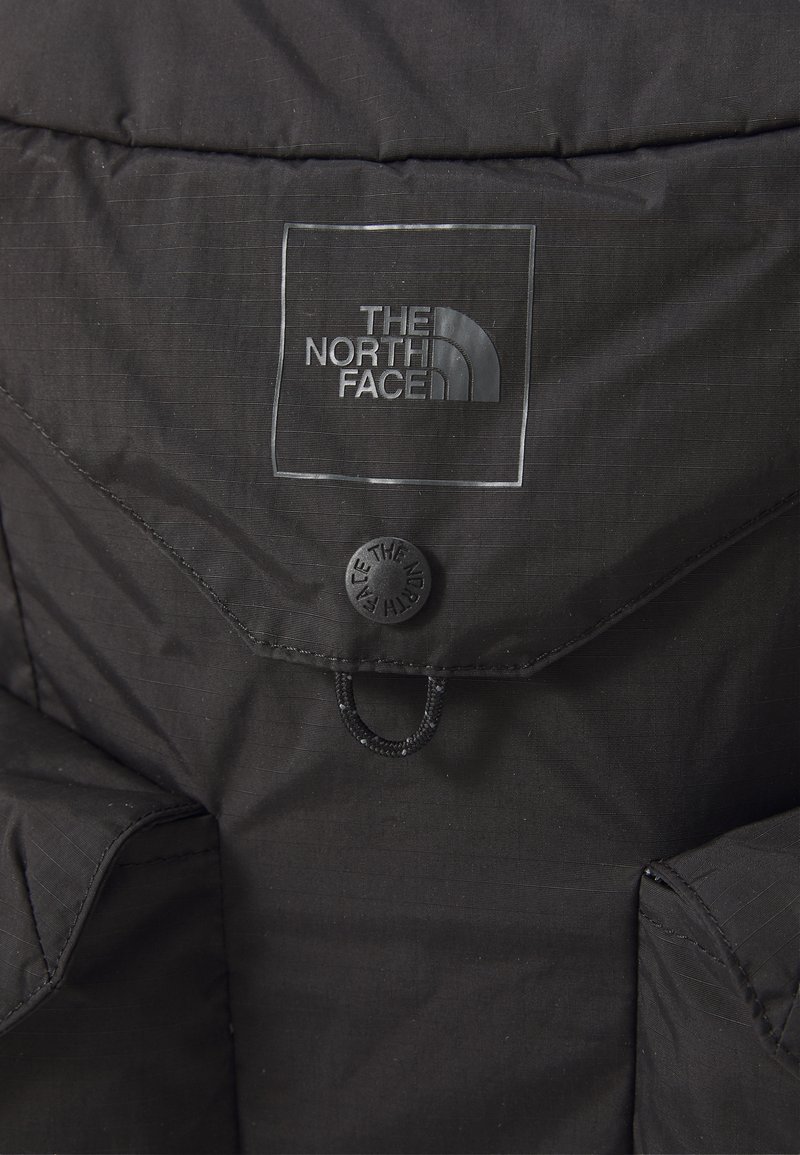 Veste isolante noire présentant un patch logo proéminent, une fermeture à boutons et un tissu texturé avec un léger motif linéaire.