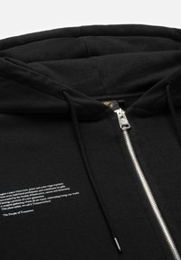 Zwarte zip-up hoodie met een zilveren ritssluiting, trekkoorden en witte tekst op de voorkant. Zachte textuur en een comfortabele pasvorm.