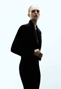 Blazer noir ajusté avec des épaules structurées, port�é par-dessus un haut noir. L'arrière-plan est une lumière douce et lumineuse, mettant en valeur la silhouette.