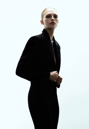 Blazer noir ajusté avec des épaules structurées, porté par-dessus un haut noir. L'arrière-plan est une lumière douce et lumineuse, mettant en valeur la silhouette.