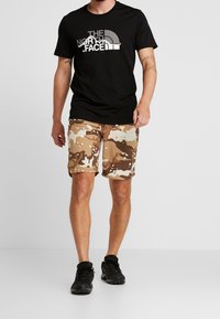 Černé tričko s krátkým rukávem s logem "The North Face", doplněné béžovými a hnědými maskáčovými šortkami a černými sportovními botami.