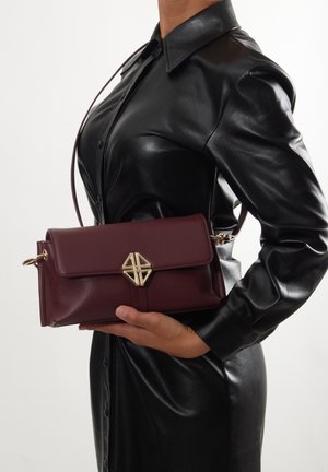 Borsa a tracolla in pelle bordeaux con patta frontale, dettagli in metallo dorato e un accento geometrico. Texture liscia con forma rettangolare.