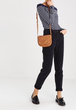 Brun mockaläder crossbody-väska med en böjd design, justerbar flätad axelrem och en metall-logotypdetalj, kombinerad med mörka jeans och en randig topp.