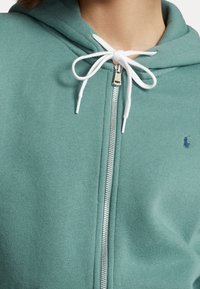 Ljust grön zip-up hoodie med en vit dragsko, med en silverfärgad dragkedja och en liten blå logotyp på vänster bröstområde. Mjuk tygkänsla.