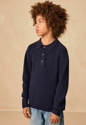 Kind mit lockigem Haar, das ein marineblaues gestricktes Langarm-Poloshirt und hellblaue Jeans trägt, schaut nach links vor einem beigen Hintergrund.
