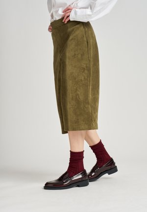 Pantalones culottes de ante verde oliva con cintura alta, combinados con calcetines de canalé rojo oscuro y mocasines burdeos pulidos. Textura suave y diseño a medida.