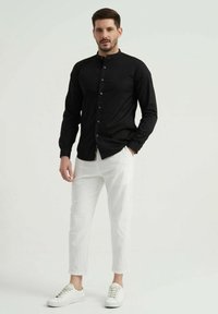 Chemise noire à manches longues avec col mandarin et fermeture à boutons, associée à un pantalon texturé blanc et des baskets blanches.