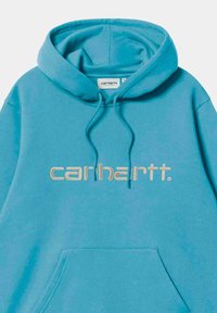 Sudadera Carhartt de color azul claro, con bolsillo frontal, capucha con cordones y logo bordado en beige en el pecho. Textura suave.