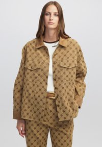 Beige gemusterte Jacke mit braunem Logo-Print, ausgestattet mit einem Kragen, zwei Brusttaschen und Knopfverschluss, kombiniert mit einem cremefarbenen Oberteil.