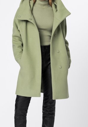 Personne portant un manteau oversize vert clair sur un pull à col roulé côtelé et un pantalon noir, mains dans les poches du manteau, sur fond blanc.
