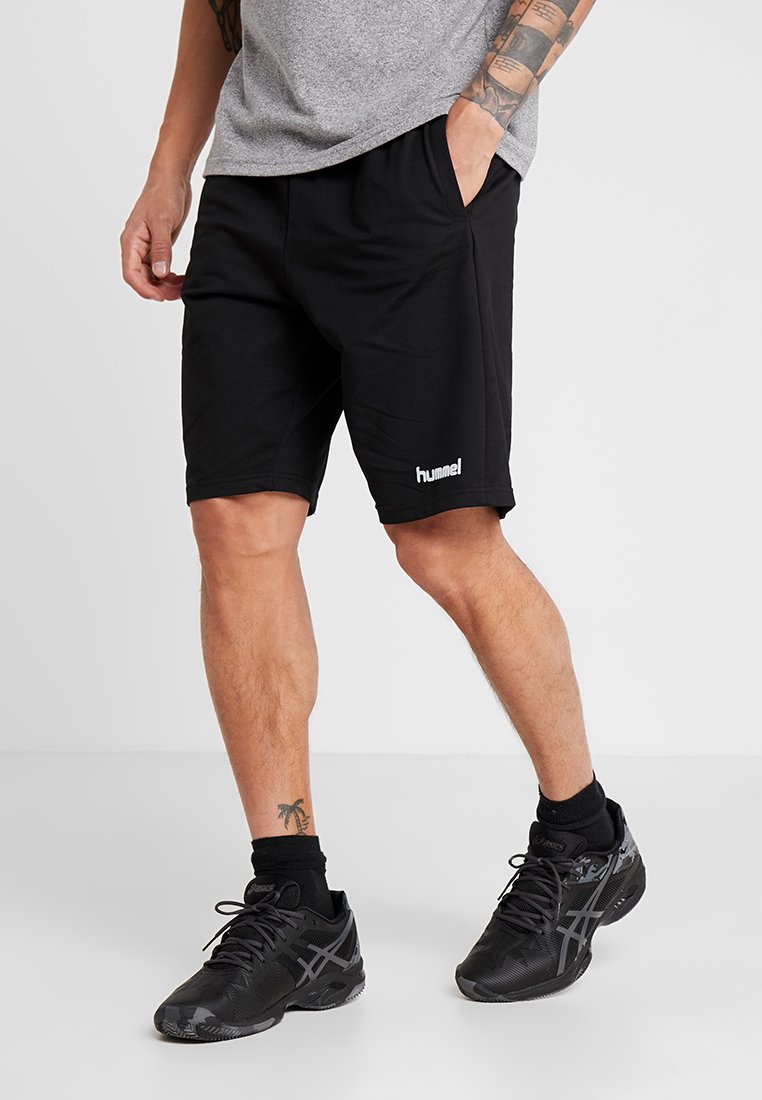 Uomo che indossa pantaloncini neri Hummel, calzini neri e scarpe da ginnastica nere, in piedi con una mano in tasca su sfondo semplice.