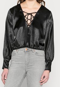 Blusa di satin nera con scollatura a V profonda e dettagli con lacci, caratterizzata da maniche lunghe e una vita aderente e arricciata. Indossata con jeans grigi.