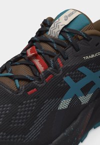 Κοντινό πλάνο μαύρου και τιρκουάζ παπουτσιού για τρέξιμο σε μονοπάτι με κόκκινες λεπτομέρειες, κορδόνια με σχέδιο και το λογότυπο "TRABUCO" στο πλάι.