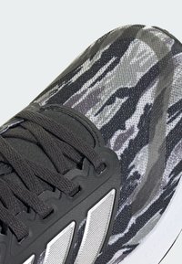 Schwarzer und grauer Sportschuh mit einer strukturierten, camouflagemusterartigen Oberseite, schwarzen Schnürsenkeln und silbernen Akzenten an der Seite. Langlebiges Material und Design.