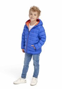 Fant nek z belimi lasmi stoji oblečen v modro puhovko z oranžnim podaljškom, svetle modre jeans hlače in bele superge pred belo ozadjem.