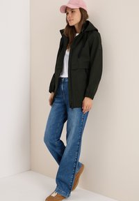 Veste à capuche vert foncé avec fermeture éclair à l'avant, deux poches latérales, assortie à un jean flare bleu clair et des baskets marron, sur un fond neutre.