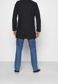 Černý saka na míru s jediným rozparkem, doplněné modrými džínami s rovnými nohavicemi a černými teniskami. Důraz na střih a čistý design.