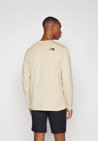 Beige langermet skjorte laget av myk stoff, med rund hals og en liten svart logo på øvre rygg. Kombinert med mørke shorts.