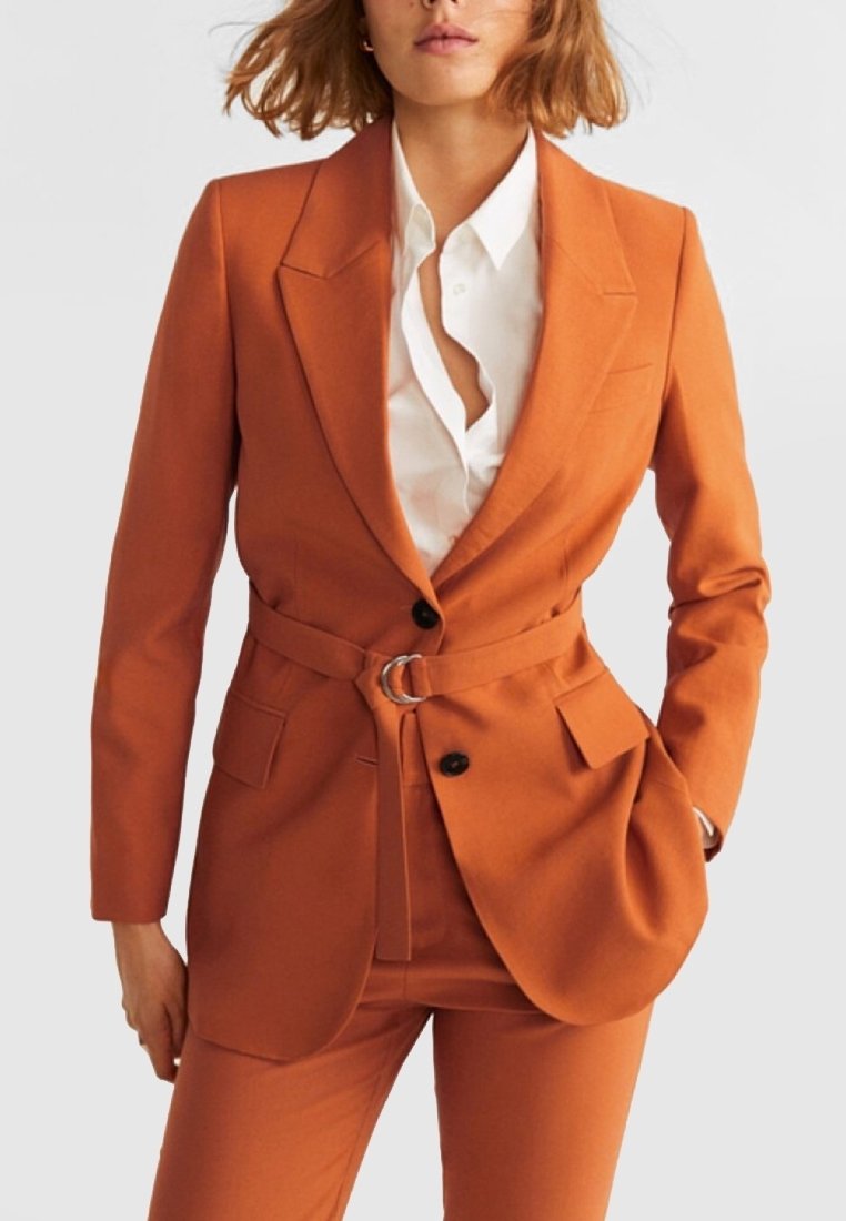 Blazer orange sur mesure avec une silhouette ajustée, fermeture à un bouton, ceinture avec anneau en métal, et deux poches avant, associé à une chemise blanche.