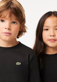 Due magliette a maniche lunghe nere su due bambini, con un piccolo logo ricamato di un coccodrillo verde e bianco sul lato sinistro di ciascuna maglietta.