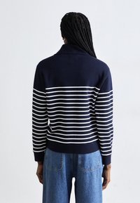 Maglione a righe blu navy e bianco con colletto alto; presenta un orlo e maniche a coste, realizzato in un morbido materiale lavorato a maglia. Jeans in denim visibili.