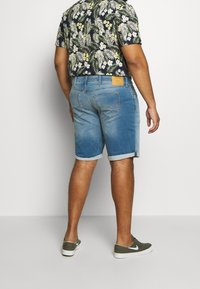 Shorts de mezclilla en azul claro con dobladillos enrollados, que presentan bolsillos traseros y una etiqueta de cuero marrón. Combinados con una camisa de estampado floral.