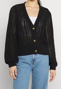Cardigan nero lavorato a maglia con scollo a V, tessuto a trama aperta, maniche lunghe a palloncino e cinque bottoni dorati sul davanti. Abbinato a jeans azzurri.