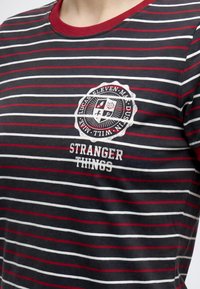 Ριγέ μαύρο, άσπρο και κόκκινο T-shirt με κόκκινο γιακά που διαθέτει κυκλικό έμβλημα και κείμενο "Stranger Things" στο στήθος.