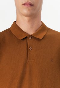 Brązowa polo z fakturowanej tkaniny, z zapięciem na guziki, krótkim kołnierzykiem oraz dyskretnym logo na piersi. Wykonana z tkaniny tkanej.
