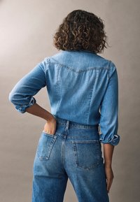 Jeansowa koszula z długimi rękawami i mankietami na guziki, w jasnoniebieskim kolorze. Połączona z wysokimi niebieskimi dżinsami, z tylnymi kieszeniami.