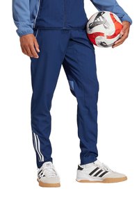 Marineblauwe sportbroek met witte strepen, gecombineerd met zwart-witte sneakers. De persoon houdt een rood-witte voetbalschoen vast.