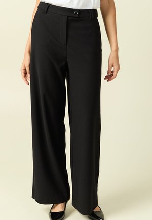 Donna con pantaloni neri a gamba larga e scarpe nere con punta affilata, in piedi davanti a uno sfondo chiaro e uniforme.