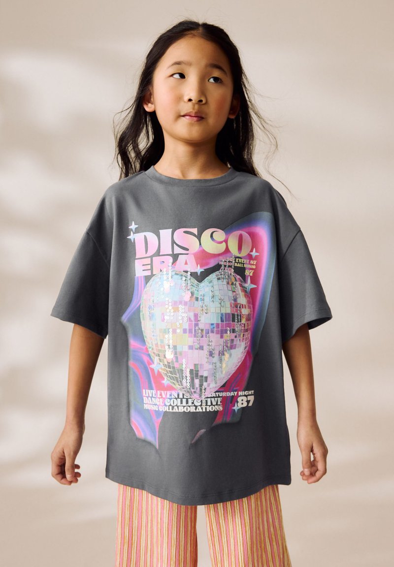 Young girl with long hair wearing a gray oversized T-shirt featuring a colorful disco heart and "DISCO ERA" text, paired with striped pants.
