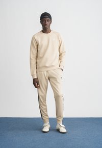 Béžová mikina s okrúhlym výstrihom a jemnou textúrou, v kombinácii so zladenými béžovými teplákmi s čiernymi bočnými pásikmi. Biele tenisky.
