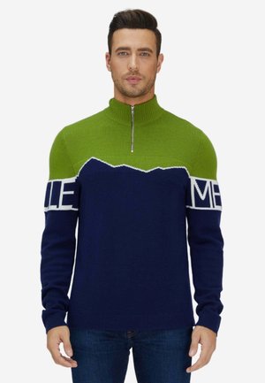 Homme portant un pull demi-fermeture éclair vert et bleu marine avec une ligne blanche en zigzag et du texte sur les manches, associé à un jean bleu, debout sur fond blanc.