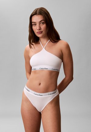 Kvinde stående med armene bag ryggen iført hvid Calvin Klein halter-bralette og matchende undertøj mod en ensfarvet grå baggrund.