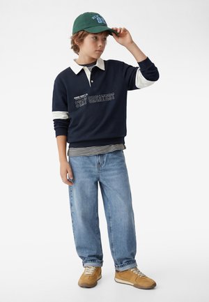 Dreng iført en marineblå sweatshirt med teksten "Stay Greatest", stribet skjorte, lyse jeans, beige sneakers og grøn kasket, der justerer kasketten med den ene hånd.