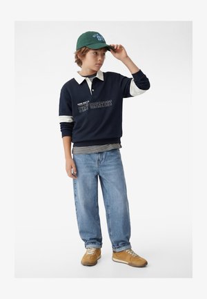 Niño con sudadera azul marino con la frase "Stay Greatest", camiseta de rayas, jeans claros, zapatillas beige y gorra verde, ajustándose la gorra con una mano.