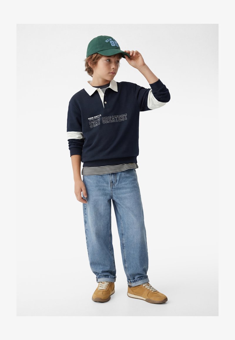 Garçon portant un sweat-shirt bleu marine avec l'inscription « Stay Greatest », une chemise rayée, un jean clair, des baskets beige et une casquette verte, ajustant la casquette d'une main.