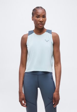 Nő világoskék ujjatlan sportos felsőben és sötétkék leggings-ben, tiszta fehér háttér előtt áll.