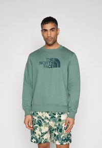 Hanorac din fleece verde cu guler rotund și logo brodat. Asortat cu pantaloni scurți cu model camuflat în nuanțe de verde și bej.