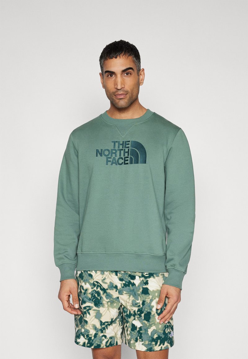 Hanorac din fleece verde cu guler rotund și logo brodat. Asortat cu pantaloni scurți cu model camuflat în nuanțe de verde și bej.