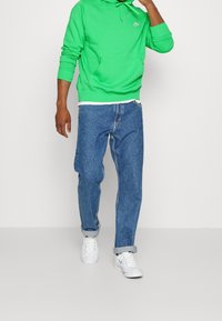 Grön huvtröja med framficka och dragsko, ihop med blå denimjeans. Vita sneakers fullända outfiten och visar på en avslappnad stil.