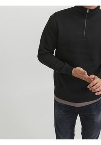 Uomo in maglione nero a maniche lunghe con zip a quarto che sistema la manica, indossando jeans scuri su uno sfondo chiaro e semplice.