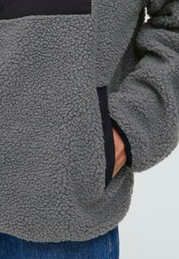 Gray fleece pullover with a textured surface, featuring a black design panel and side pockets for functionality. Soft, cozy material.