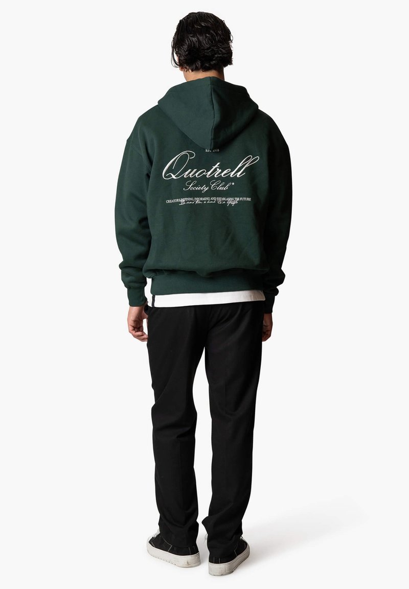 Grüne Kapuzensweatshirt mit rückseitigem Textdesign; hat gerippte Bündchen und Saum, getragen mit schwarzen Hosen und einem weißen Layered-Shirt darunter.